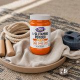 NOW Foods L-Glutamine Powder tub beside a shaker cup and juice on a kitchen counter
