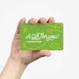 A hand holding a green gift card for The Healthy Place. The card features white cursive text reading "A Gift for You!" with decorative yellow leaf and swirl designs. A simple and clean background emphasizes the card.