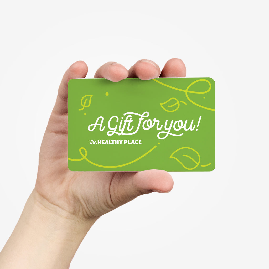 A hand holding a green gift card for The Healthy Place. The card features white cursive text reading "A Gift for You!" with decorative yellow leaf and swirl designs. A simple and clean background emphasizes the card.