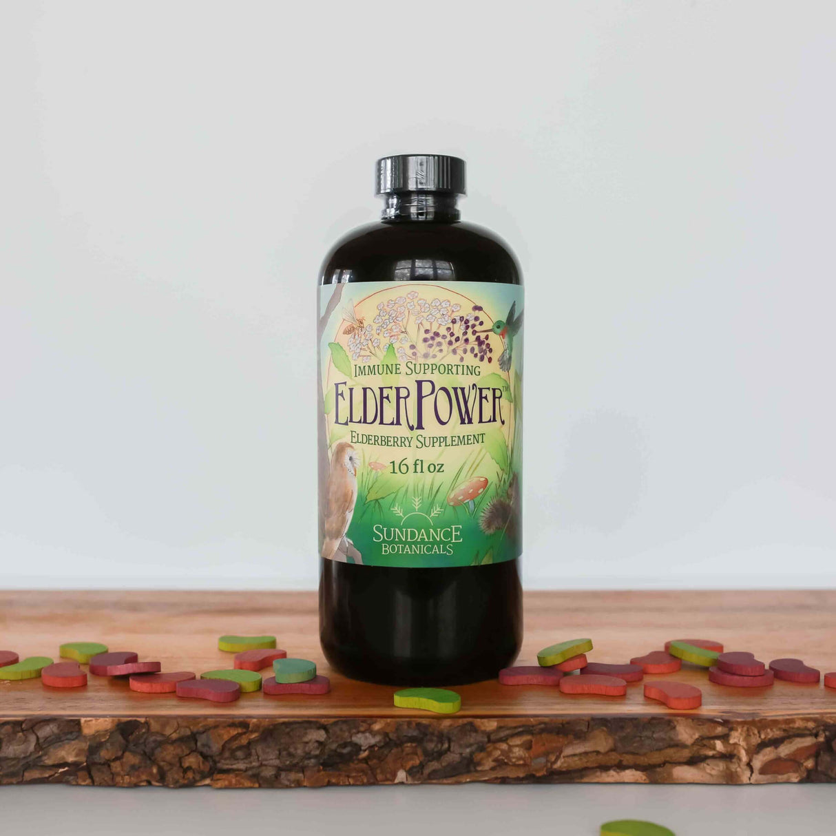 A bottle of Sundance Botanicals ElderPower Elderberry Supplement, 16 fl oz, sits on a wooden board surrounded by colorful, leaf-shaped chewable tablets. The label reads “Immune Supporting ElderPower Elderberry Supplement,” and is decorated with illustrations of elderberries, birds, mushrooms, and plants.
