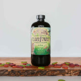 A bottle of Sundance Botanicals ElderPower Elderberry Supplement, 16 fl oz, sits on a wooden board surrounded by colorful, leaf-shaped chewable tablets. The label reads “Immune Supporting ElderPower Elderberry Supplement,” and is decorated with illustrations of elderberries, birds, mushrooms, and plants.
