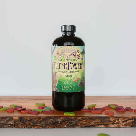 A bottle of Sundance Botanicals ElderPower Elderberry Supplement, 16 fl oz, sits on a wooden board surrounded by colorful, leaf-shaped chewable tablets. The label reads “Immune Supporting ElderPower Elderberry Supplement,” and is decorated with illustrations of elderberries, birds, mushrooms, and plants.
