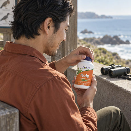 NOW Foods Kelp 325 mcg bottle held by a man at a coastal lookout, binoculars on the wall beside him