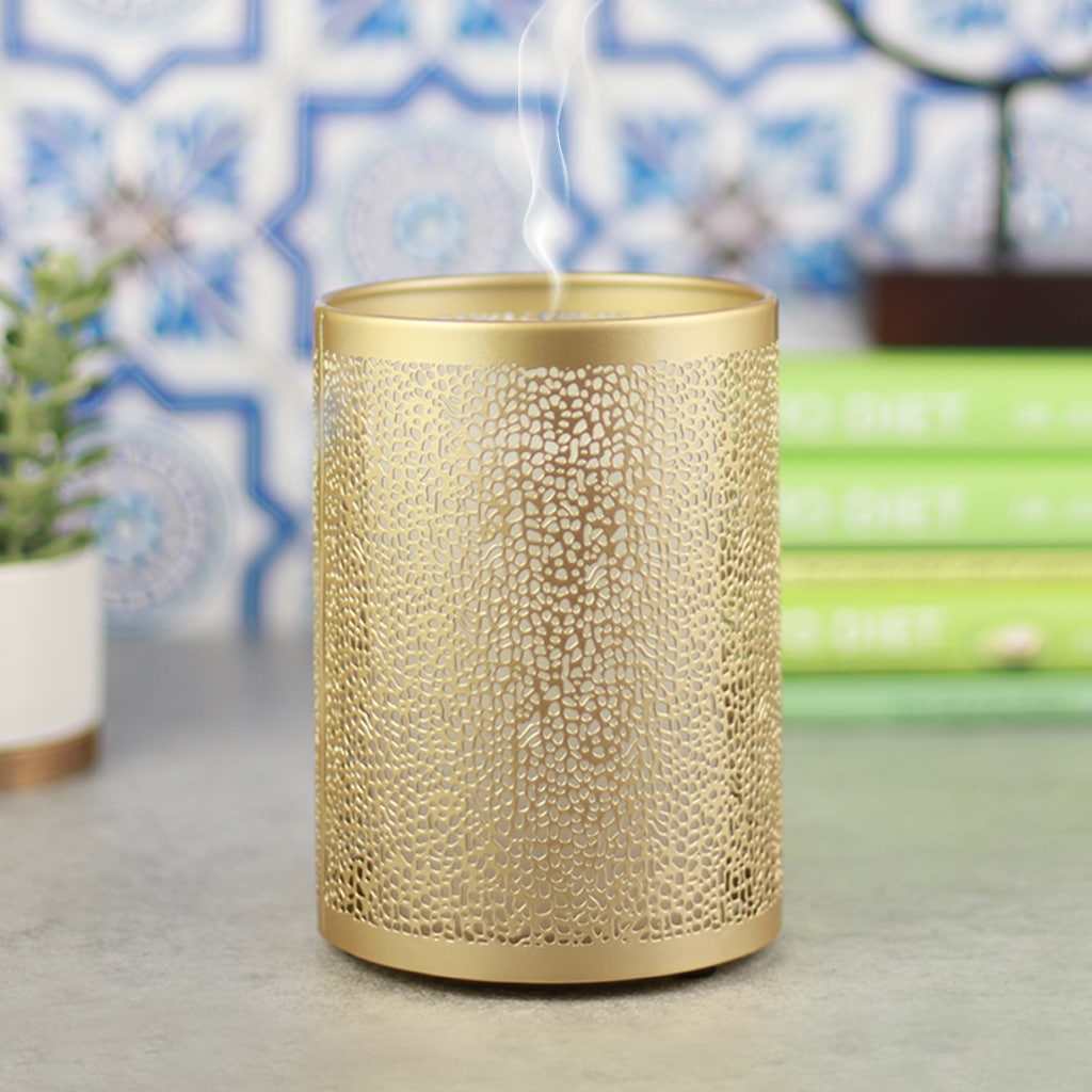 A stylish gold essential oil diffuser with intricate patterns releasing a wisp of vapor. It is set against a decorative background with plants and books.